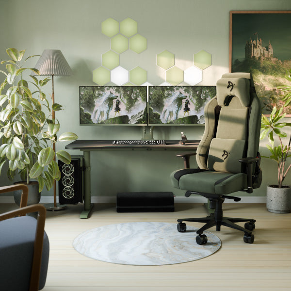 Modern home office with a gaming chair, dual monitors, green plants, hexagonal wall lights, a round rug, and a framed picture of a castle on green walls. A desk lamp sits on the left side.